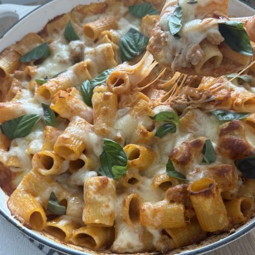 Overhead view of baked rigatoni and Italian sausage in a round pan, topped with melted mozzarella, tomato sauce, and fresh basil leaves.