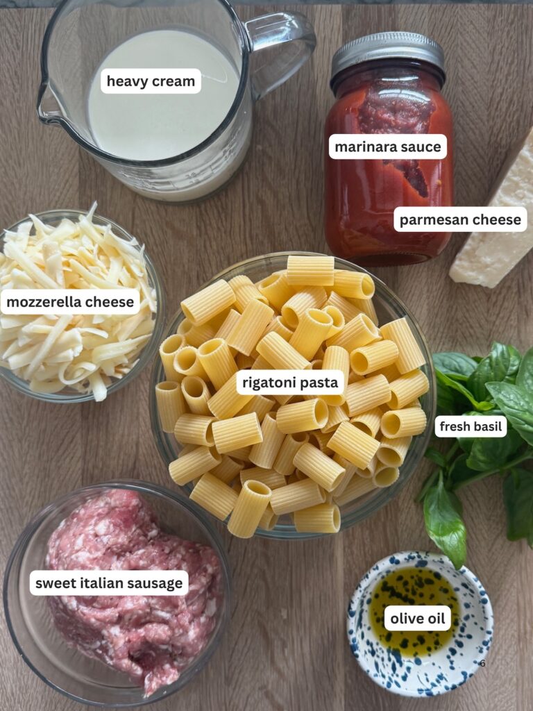 Overhead image of ingredients for baked rigatoni and Italian sausage: rigatoni pasta, marinara sauce, heavy cream, mozzarella, Parmesan cheese, sweet Italian sausage, olive oil, and fresh basil on a wooden board.