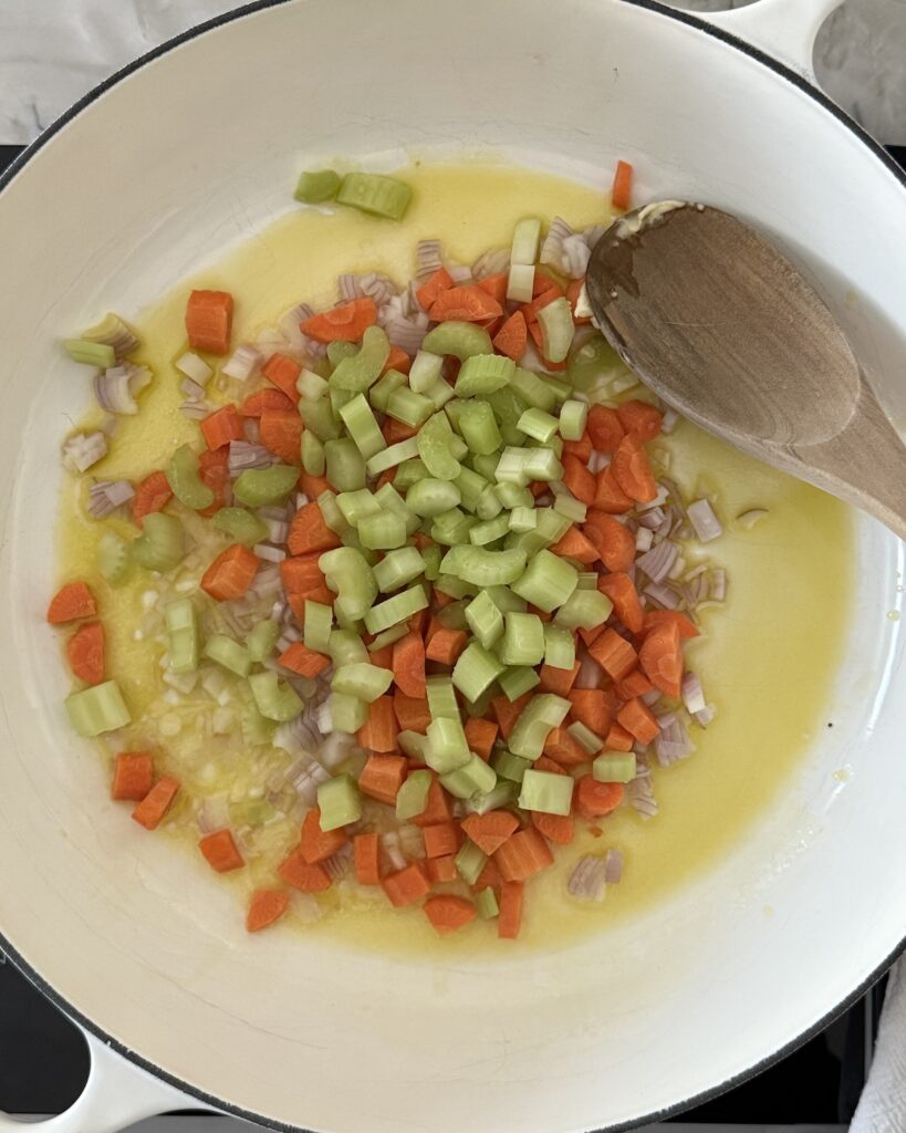 Diced carrots, celery, and shallot sautéing in butter in a white Dutch oven as the base for Italian chicken gnocchi soup.