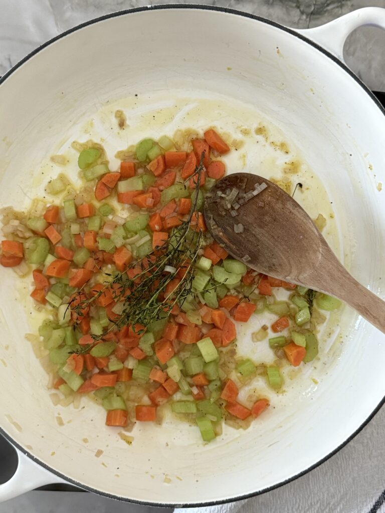 Diced carrots, celery, and shallot, and fresh thyme sprigs, sautéing in butter in a white Dutch oven as the base for Italian chicken gnocchi soup.