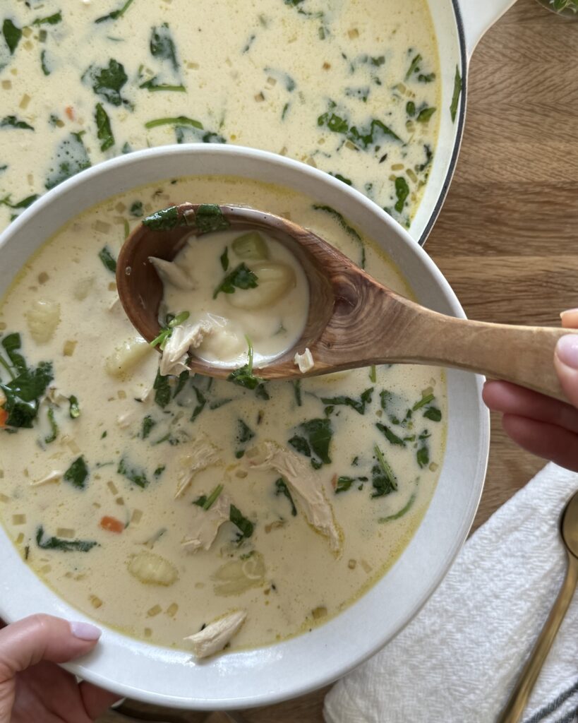 Italian chicken gnocchi soup in a white Dutch oven with creamy broth, shredded chicken, gnocchi, and baby kale, styled on a wooden board with a wooden spoon.