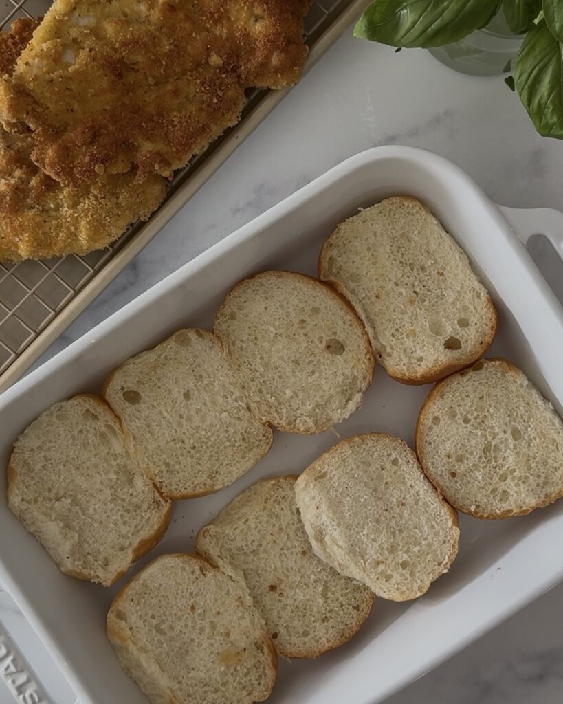 Bottom halves of slider rolls lined in a white baking dish—the base for Chicken Parm Sliders.