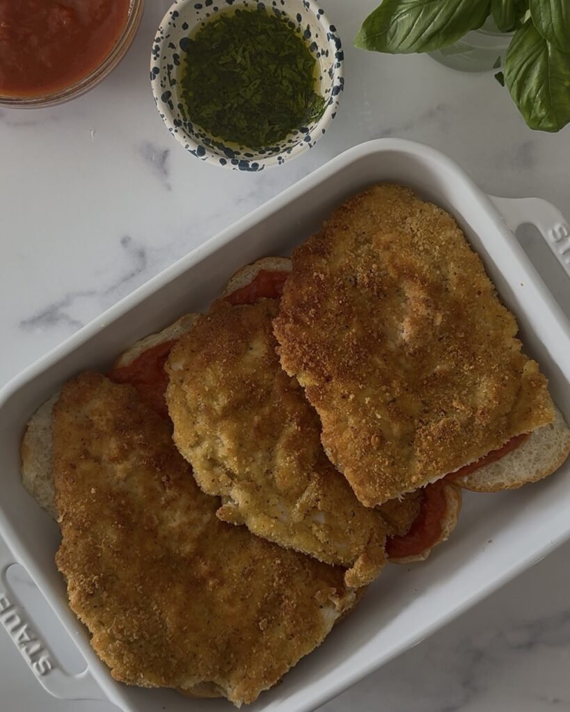 Cooked breaded chicken cutlets layered in a baking dish for chicken parmesan sliders, ready for sauce and cheese.