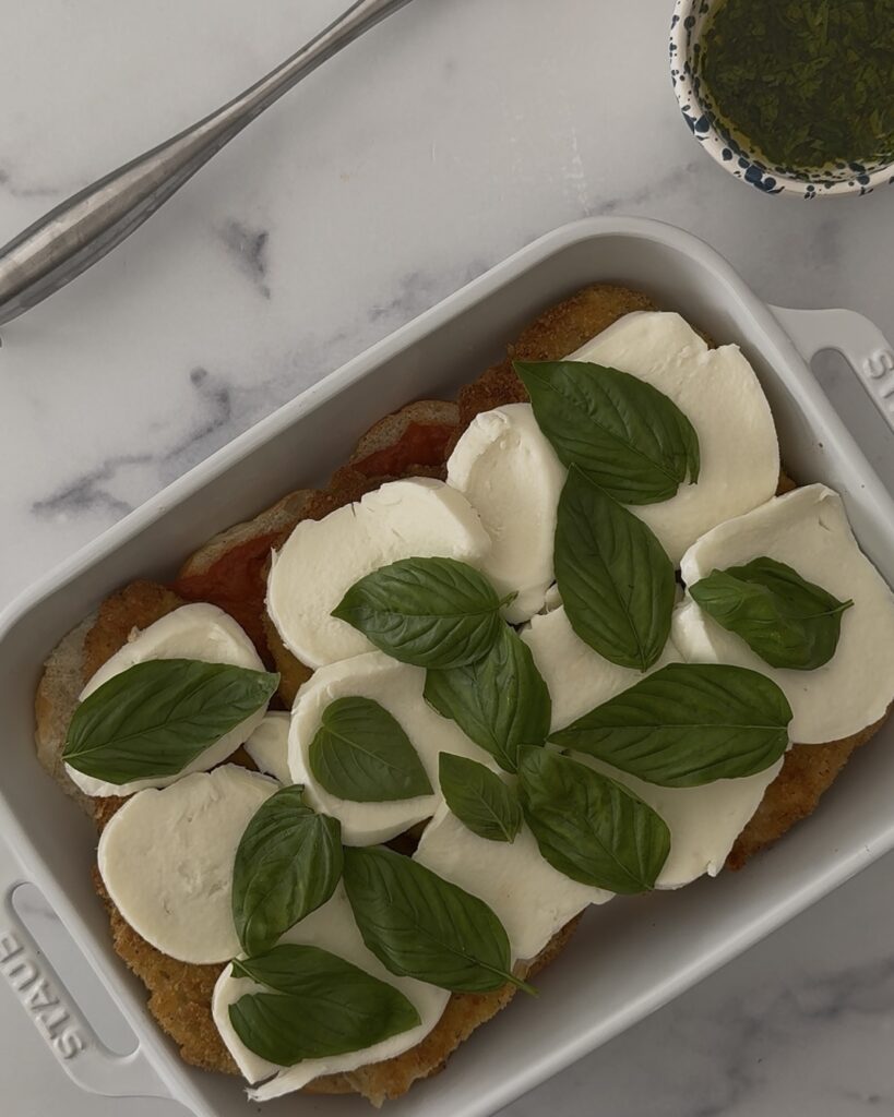 Assembly step—fresh mozzarella and basil layered over the chicken for chicken parmesan sliders in a baking dish.