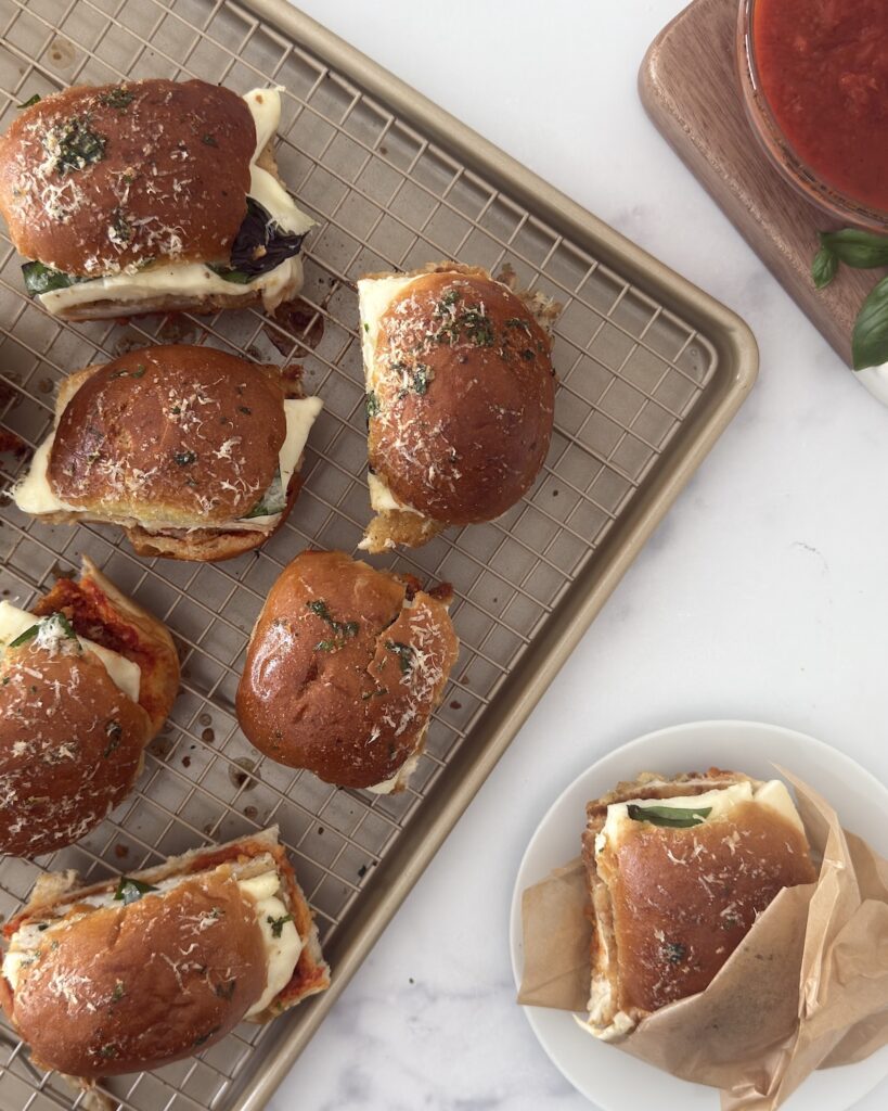 Baked chicken parmesan sliders on a cooling rack, with one served on parchment beside a bowl of marinara.