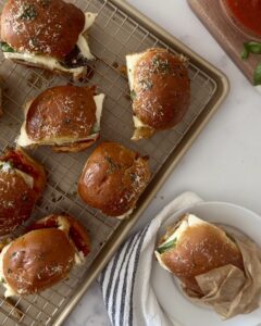 Baked chicken parmesan sliders on a cooling rack, with one served on parchment beside a bowl of ma