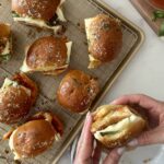 Hand holding a chicken parmesan slider with melty cheese; baked sliders on a cooling rack with a bowl of marinara nearby.