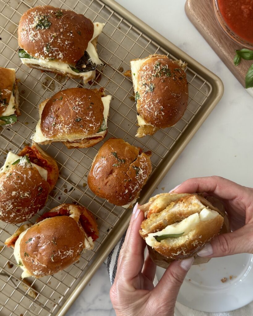 Hand holding a chicken parmesan slider with melty cheese; baked sliders on a cooling rack with a bowl of marinara nearby.