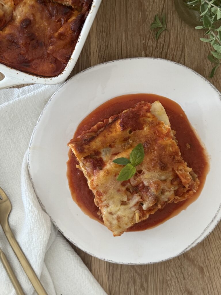 Slice of homemade meatball lasagna served on a bed of marinara sauce, garnished with fresh basil on a white plate.