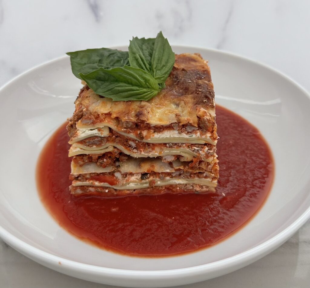 Tall slice of homemade meatball lasagna layered with ricotta and sauce, served on a bed of marinara and topped with fresh basil.