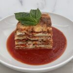 Tall slice of homemade meatball lasagna layered with ricotta and sauce, served on a bed of marinara and topped with fresh basil.