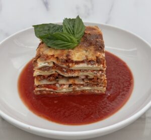Tall slice of homemade meatball lasagna layered with ricotta and sauce, served on a bed of marinara and topped with fresh basil.