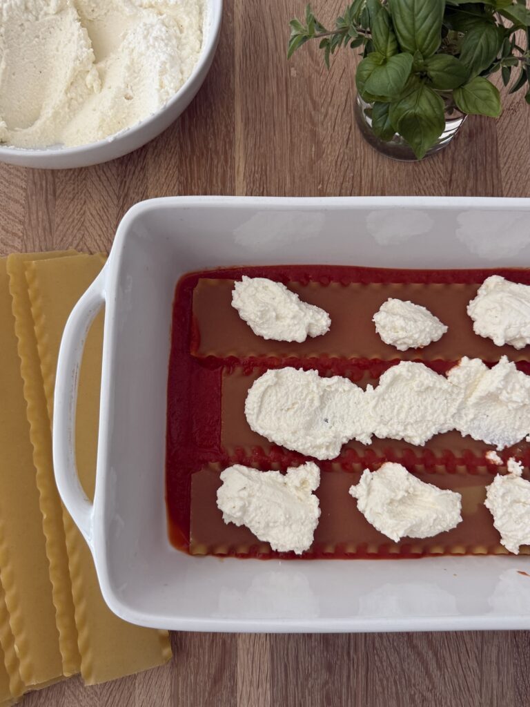 First layer of meatball lasagna with smooth marinara and dollops of ricotta in a baking dish, with no-boil lasagna noodles and grated cheese ready to add.