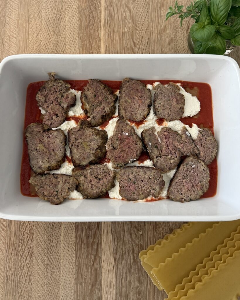 First layer of homemade meatball lasagna in a baking dish with marinara, ricotta, and sliced meatballs, with no-boil lasagna noodles ready to layer.