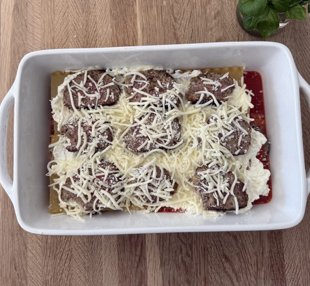 Layer of homemade meatball lasagna in a white baking dish, with ricotta, marinara, and shredded cheese ready to bake.