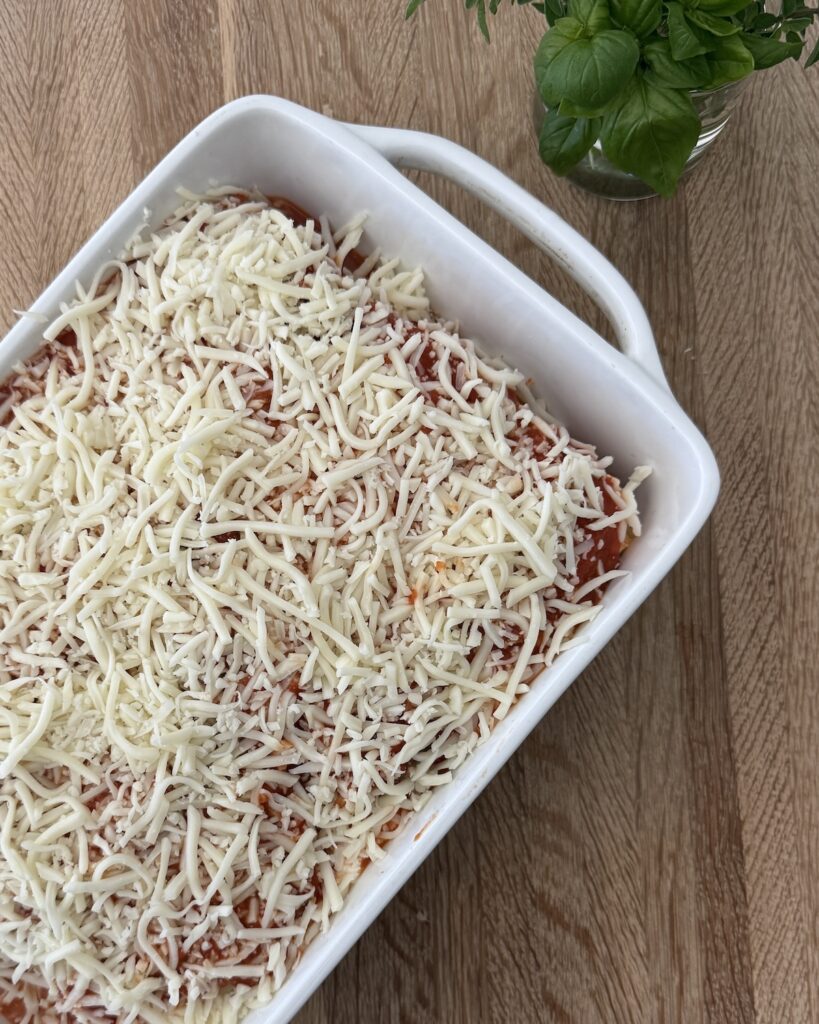 Unbaked homemade meatball lasagna in a white baking dish, topped with shredded mozzarella and ready to bake with fresh basil nearby.