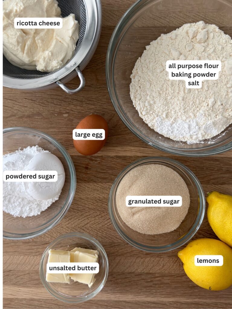 Lemon ricotta cookie ingredients on a countertop: ricotta cheese, flour, baking powder, salt, egg, granulated sugar, powdered sugar, unsalted butter, and fresh lemons.