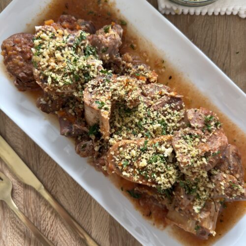 Slow cooker osso buco arranged on a serving platter with braising sauce and topped with breadcrumb gremolata, with extra gremolata in a small bowl on the side.