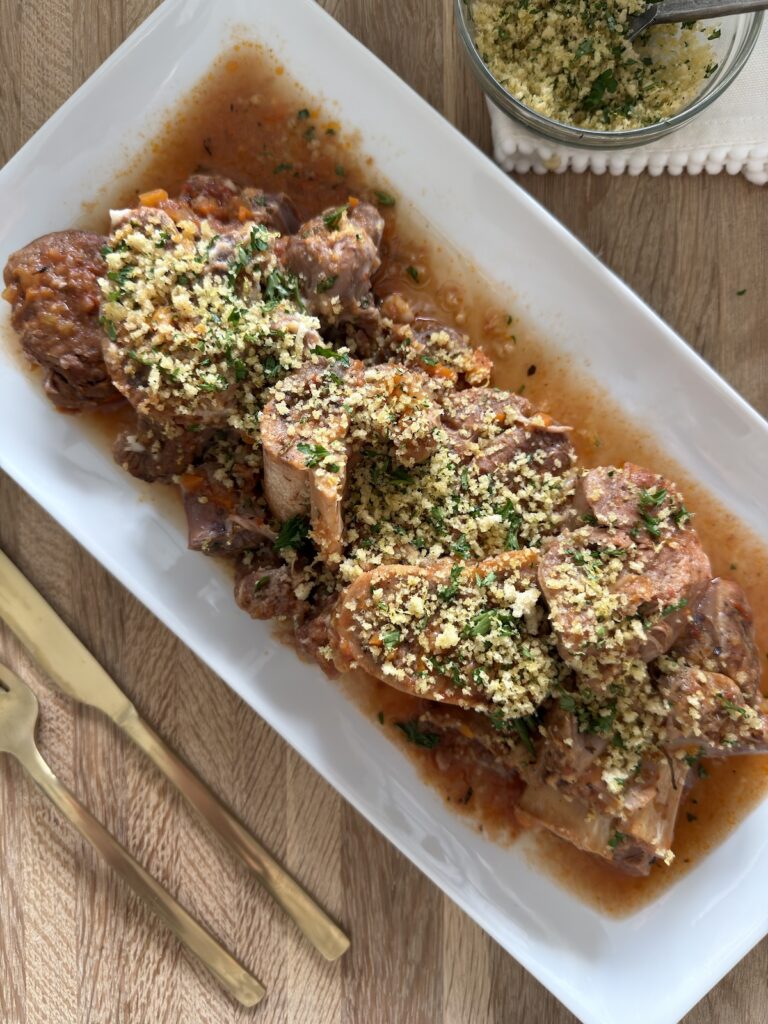 Slow cooker osso buco arranged on a serving platter with braising sauce and topped with breadcrumb gremolata, with extra gremolata in a small bowl on the side.