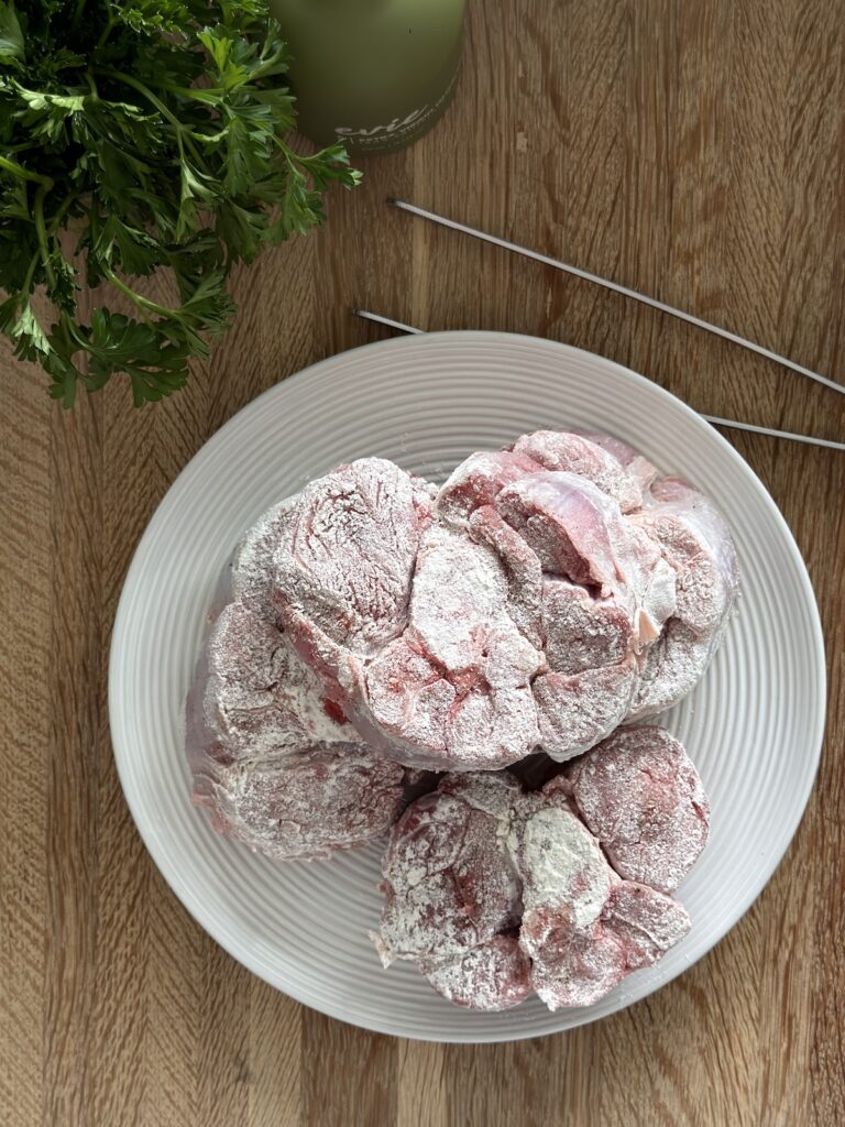 Veal shanks are floured on a white plate and ready to be seared with fresh parsley in the background.