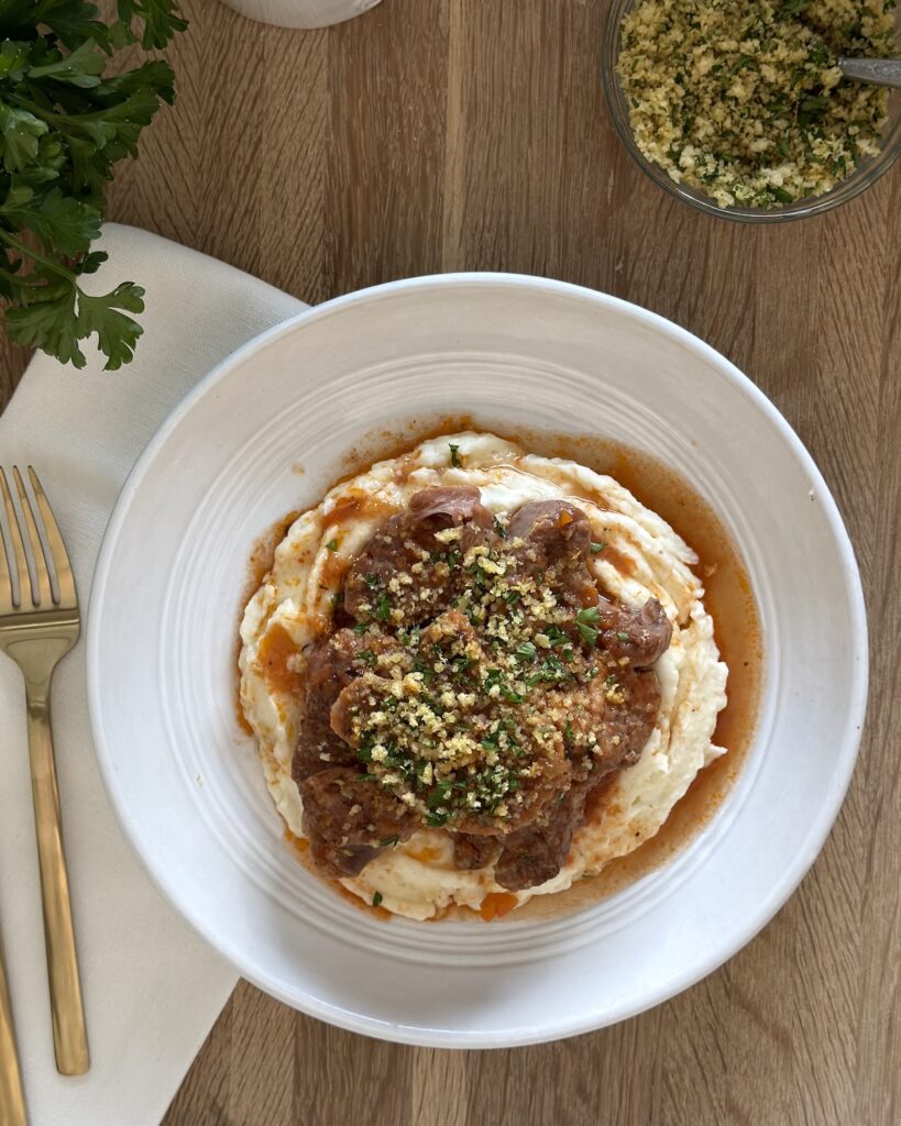 Plated slow cooker osso buco served over mashed potatoes and topped with breadcrumb gremolata, with extra gremolata and fresh parsley on the side.