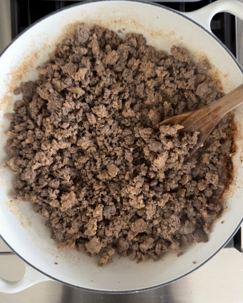 Overhead view of browned ground beef and Italian sausage in a white Dutch oven with a wooden spoon, the first step for baked bolognese pasta