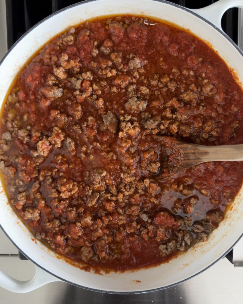 Overhead view of ground beef and Italian sausage bolognese sauce simmering in a white Dutch oven with a wooden spoon, the base for baked bolognese pasta
