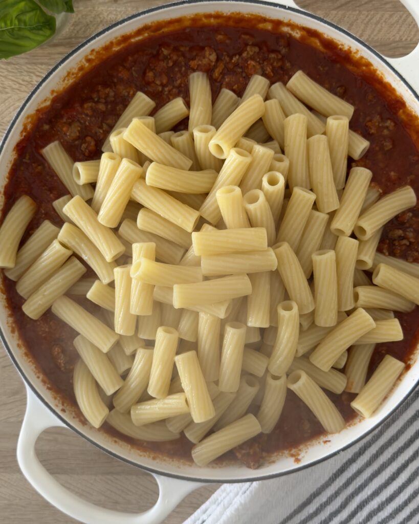 Overhead view of rigatoni pasta added to bolognese sauce in a white Dutch oven, ready to be mixed for baked bolognese pasta