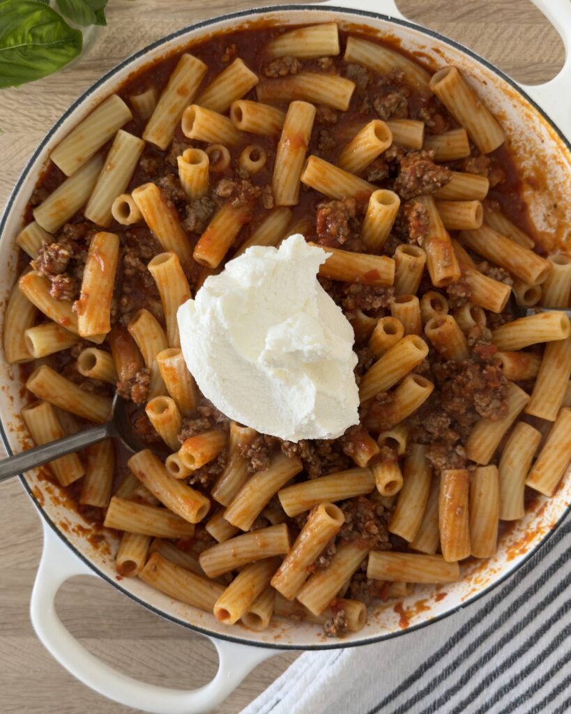 Overhead view of  bolognese pasta in a white Dutch oven with a large dollop of ricotta cheese in the center, ready to be mixed into the sauce