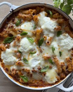 Overhead view of baked rigatoni bolognese pasta in a white pan topped with golden melted mozzarella and fresh basil