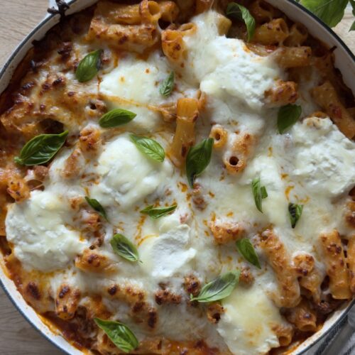 Overhead view of baked rigatoni bolognese pasta in a white pan topped with golden melted mozzarella and fresh basil
