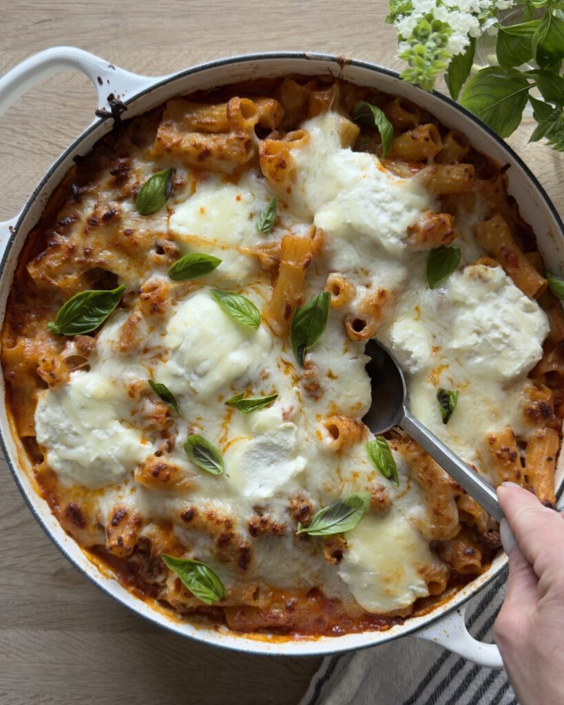 Overhead view of baked bolognese pasta in a white pan topped with melted mozzarella and basil, with a hand holding the handle for serving