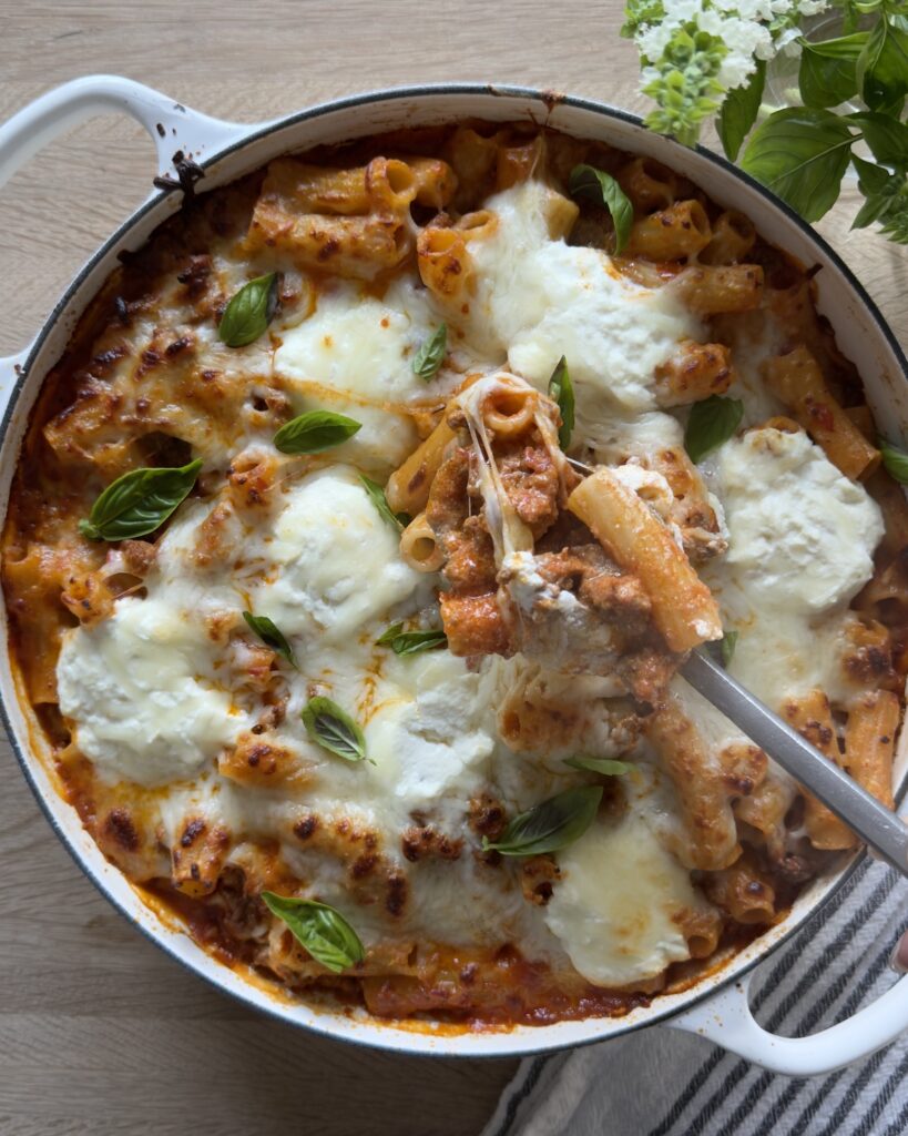 Overhead view of bakedi bolognese pasta with melted mozzarella and basil, with a spoon lifting a cheesy portion from the pan