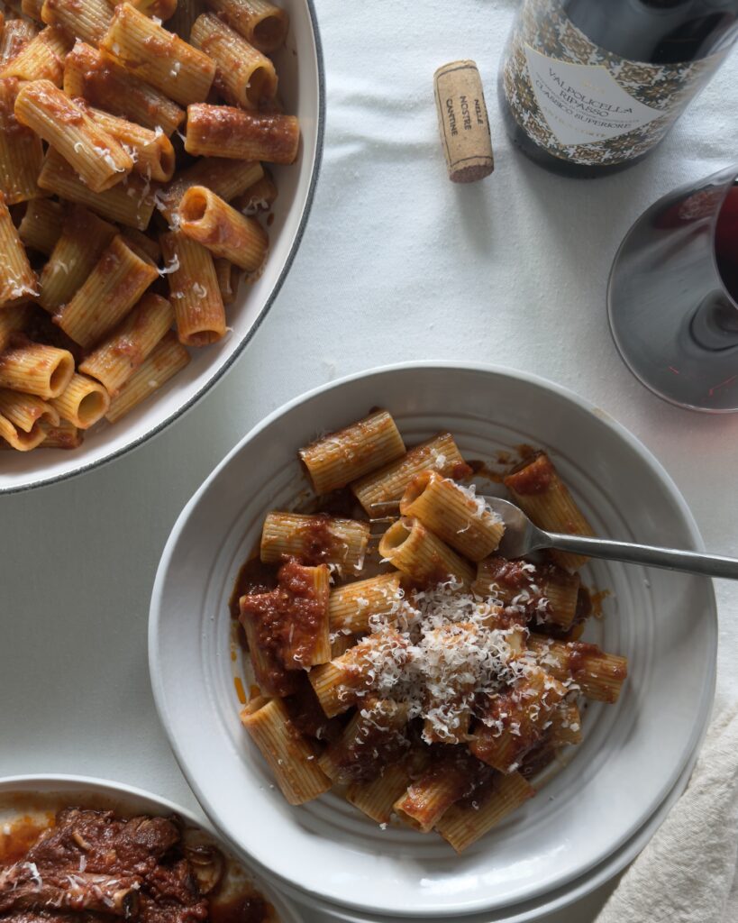 Rigatoni tossed with Italian Sunday sauce made with braised short ribs, finished with grated Parmesan cheese ona white tablecloth in white bowls, ready to be served.