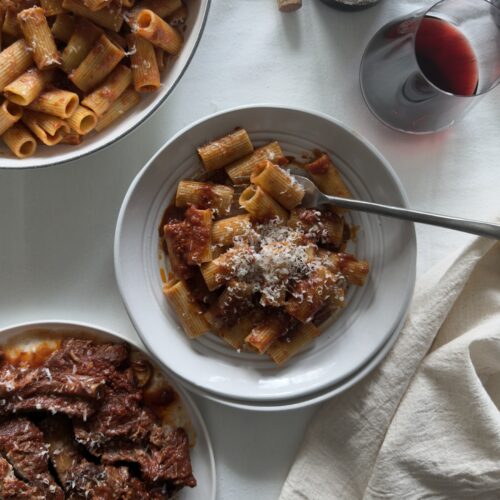 Rigatoni tossed with Italian Sunday sauce made with braised short ribs, finished with grated Parmesan cheese ona white tablecloth in white bowls, ready to be served.