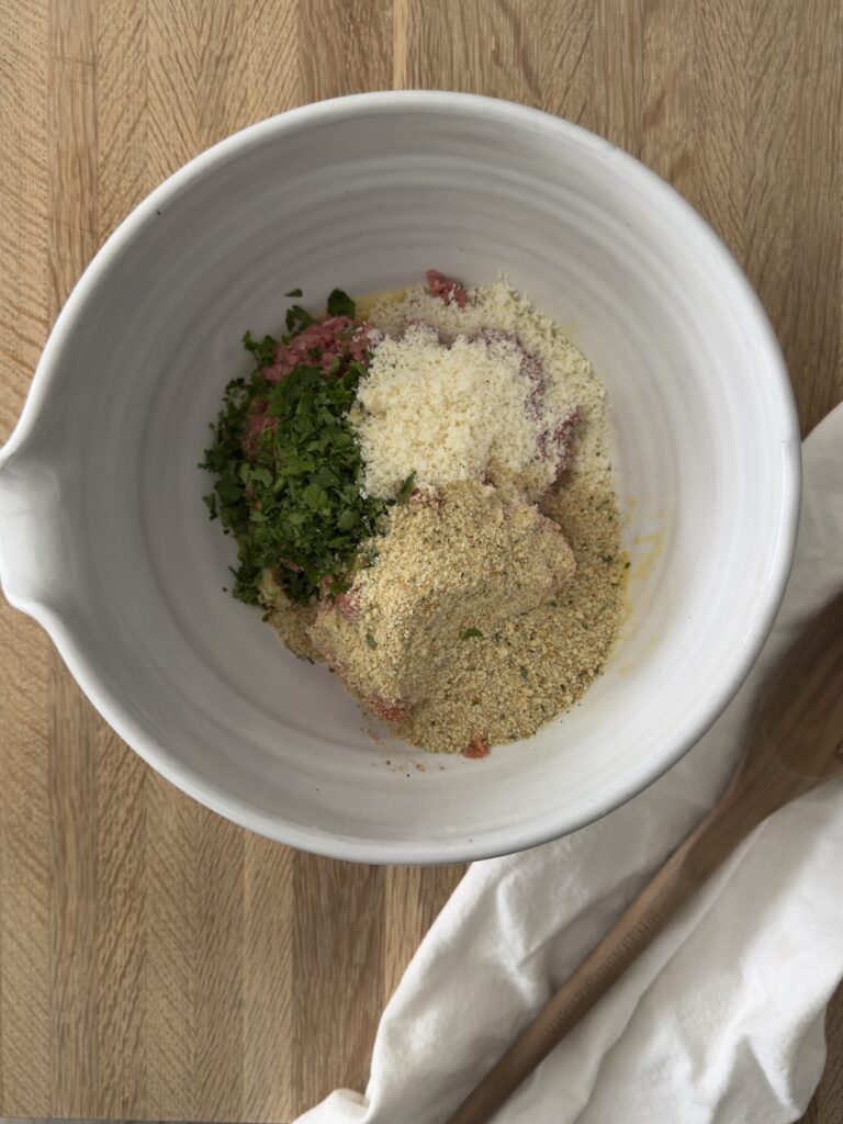 Italian wedding soup meatball ingredients in a mixing bowl with ground meat, breadcrumbs, grated Parmesan cheese, and fresh herbs before mixing.