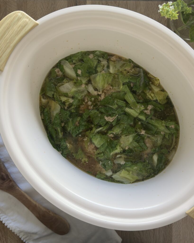 Italian wedding soup is simmering in a white crock pot with meatballs, leafy greens, and broth cooking together.