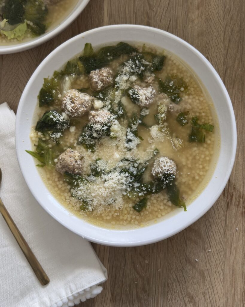 Overhead view of Italian wedding soup with meatballs, acini di pepe pasta, leafy greens, and grated Parmesan cheese in a white bowl on a wooden table.