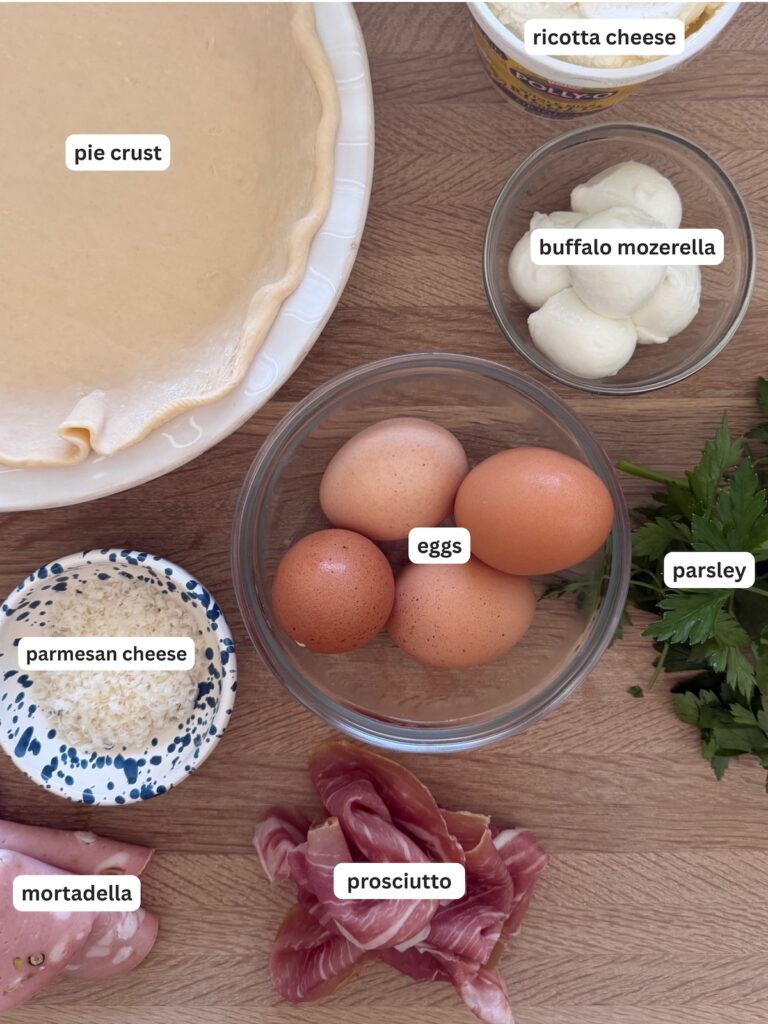 Italian Easter Pie ingredients including pie crust, ricotta cheese, buffalo mozzarella, eggs, prosciutto, mortadella, parsley, and kosher salt arranged on a wooden surface
