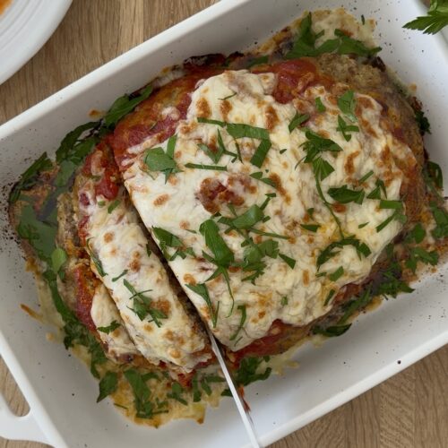 Italian meatloaf topped with melted mozzarella and fresh parsley being sliced in a white baking dish