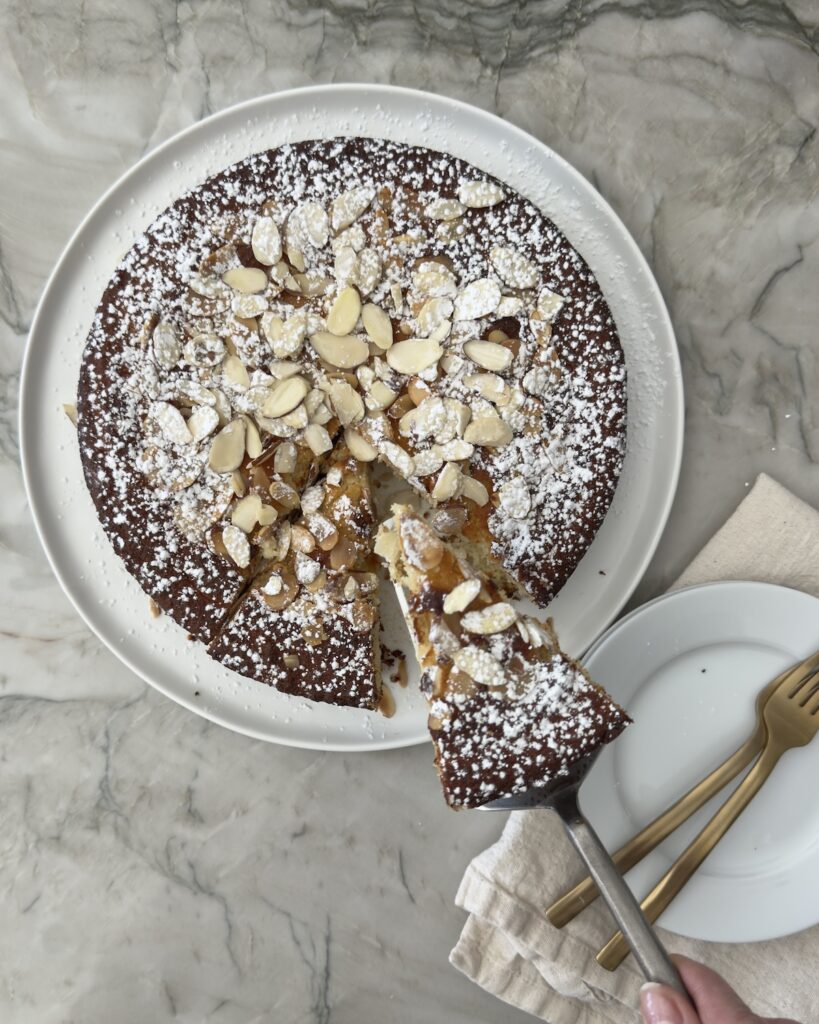 Slice of Italian almond ricotta cake lifted from the pan, showing the moist interior, topped with powdered sugar and sliced almonds on a marble surface
