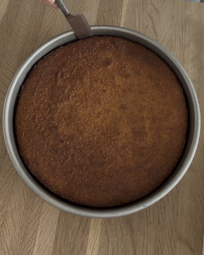 Freshly baked almond ricotta cake cooling in a round cake pan with a golden brown top on a wooden surface