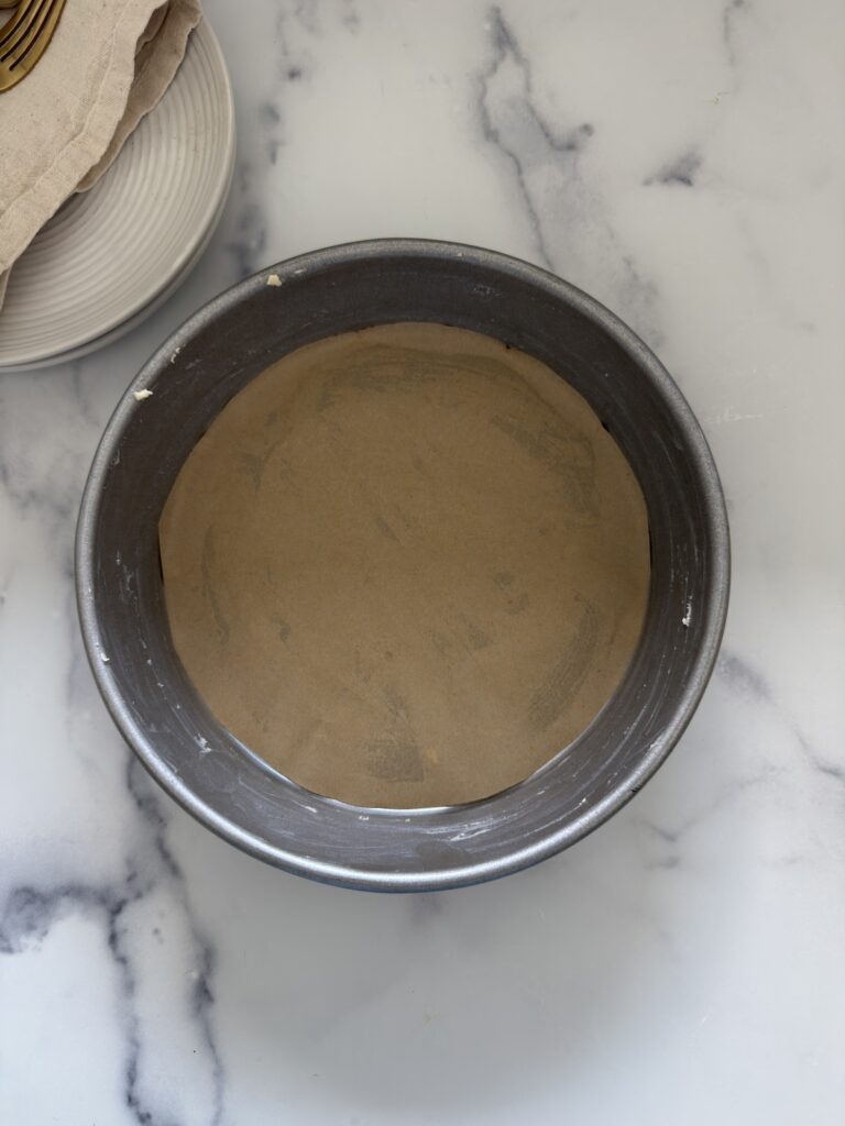 Butter-greased round cake pan lined with parchment paper prepared for baking almond ricotta cake on a marble countertop