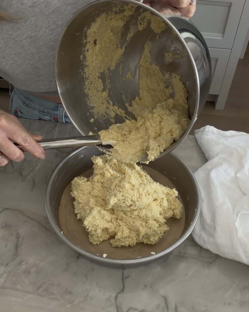 Almond ricotta cake batter being scraped from a mixing bowl into a parchment-lined round cake pan before baking