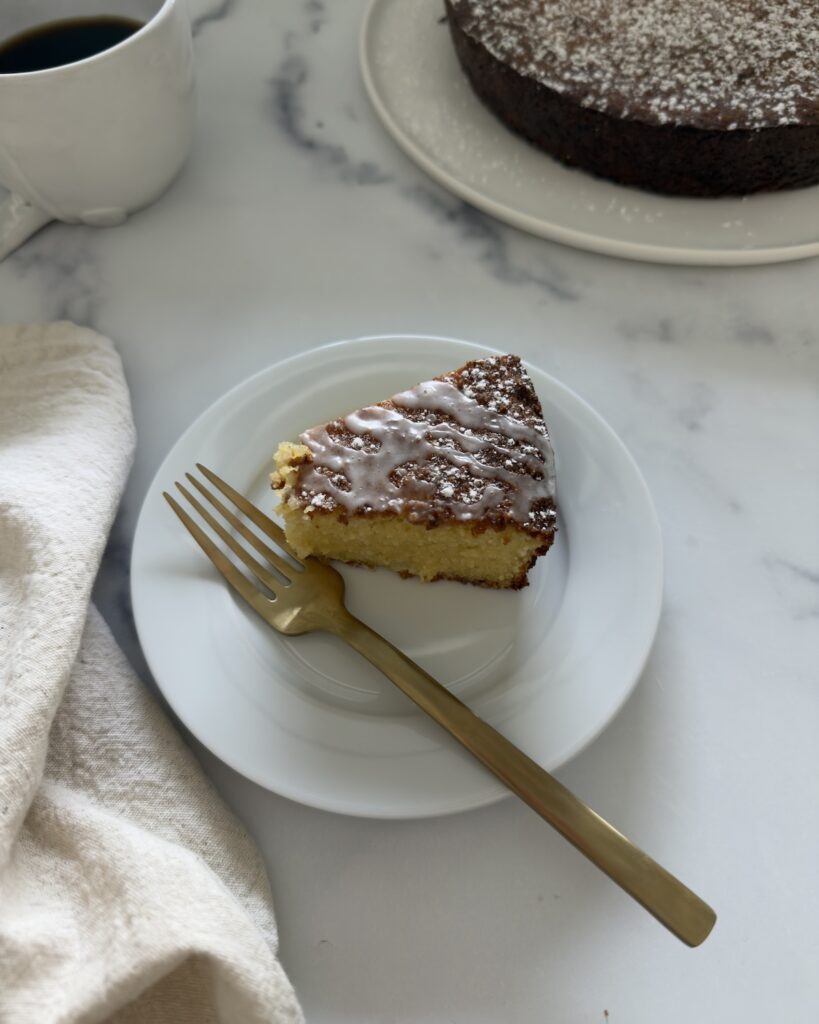 Slice of almond ricotta cake dusted with powdered sugar served on a white plate with a fork, with the whole cake in the background on a marble surface