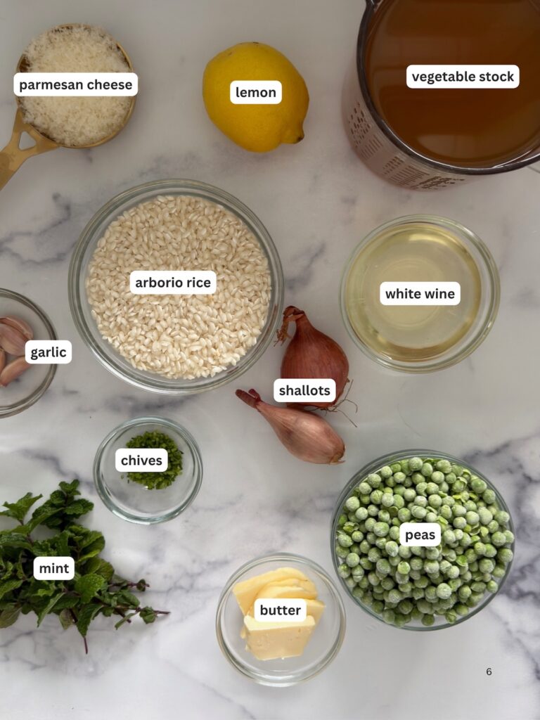 Ingredients for pea risotto including arborio rice, frozen peas, Parmesan cheese, shallot, garlic, lemon, white wine, vegetable stock, butter, mint, and chives arranged on marble surface.