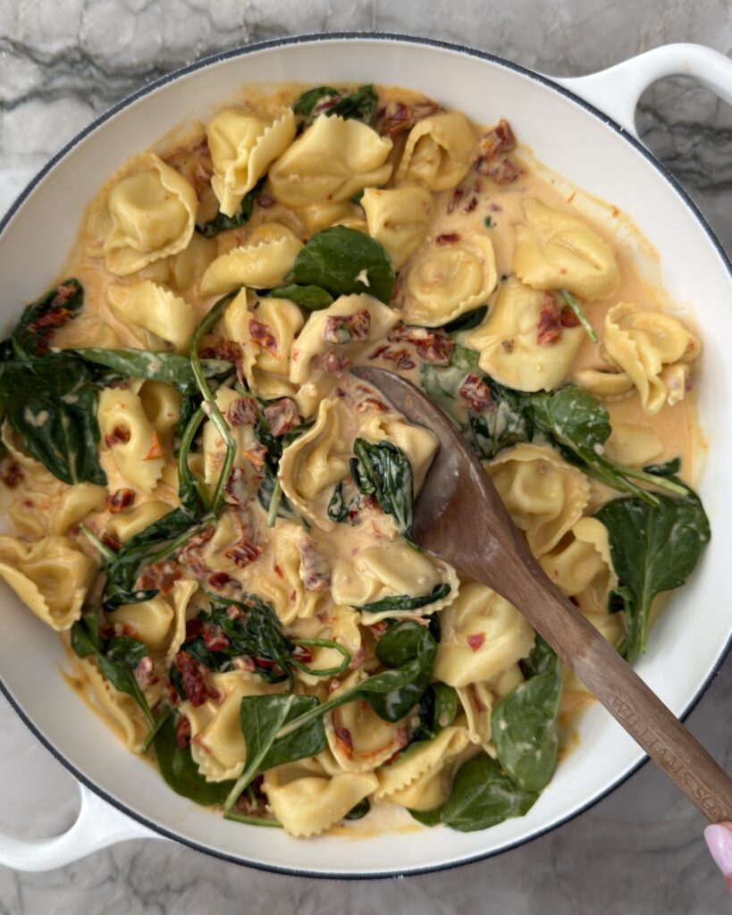 Creamy sun-dried tomato tortellini with spinach being stirred in a white braiser