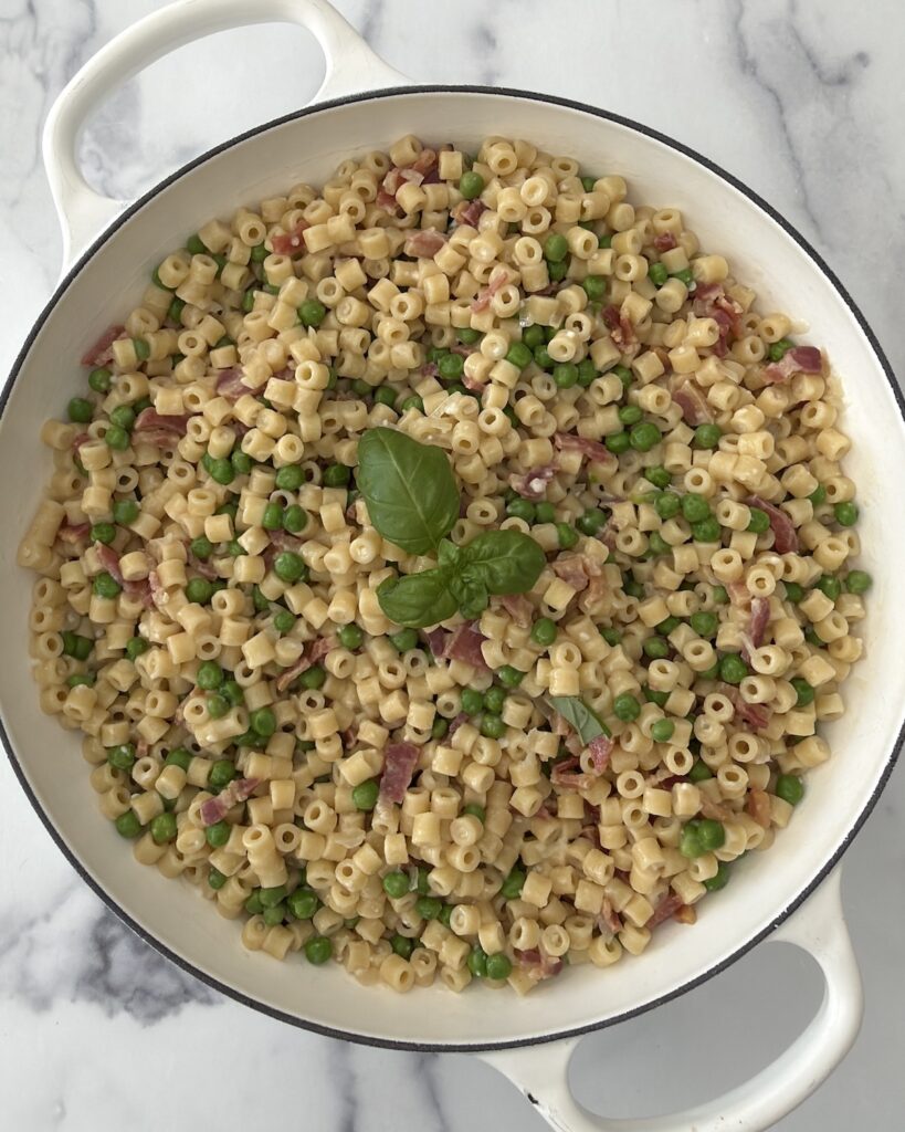 Creamy pasta e piselli with ditalini, sweet peas, crispy pancetta, and fresh basil served in a white skillet.
