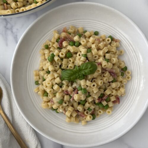 Bowl of creamy pasta e piselli with ditalini, peas, pancetta, and fresh basil served on a marble countertop.
