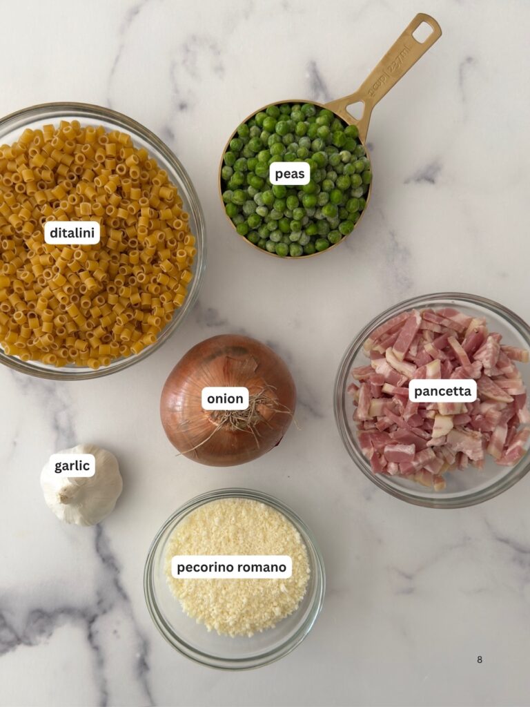 Ingredients to make pasta e Pirelli, including Ditalini pasta, peas, onion, garlic, pecorino, and pancetta, are displayed on a marble countertop.